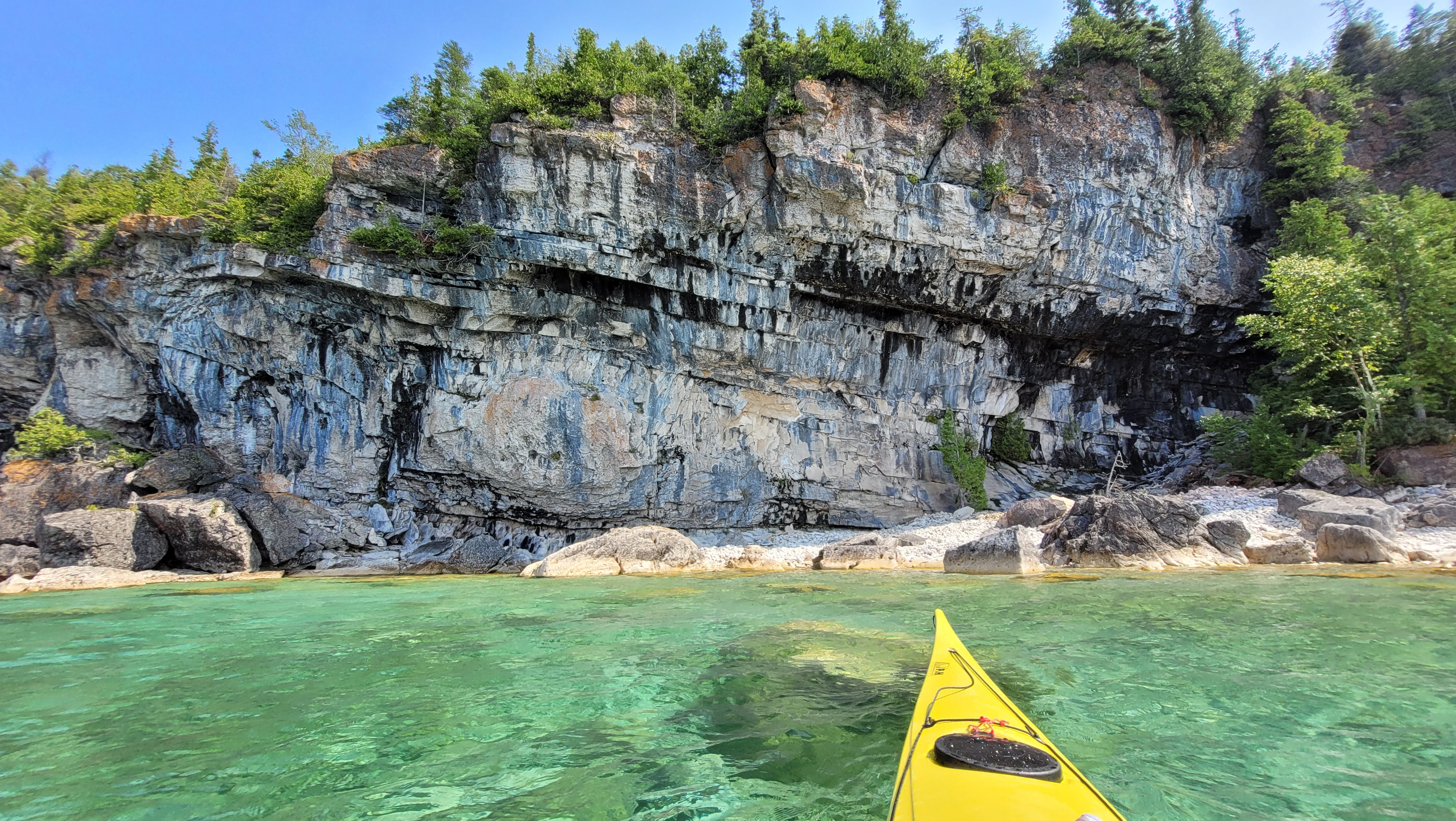 A white limestone cliffs with grays and blacks and trees on top, and clear turqouise water below in Driftwood Cove in Bruce PEninsula National Park