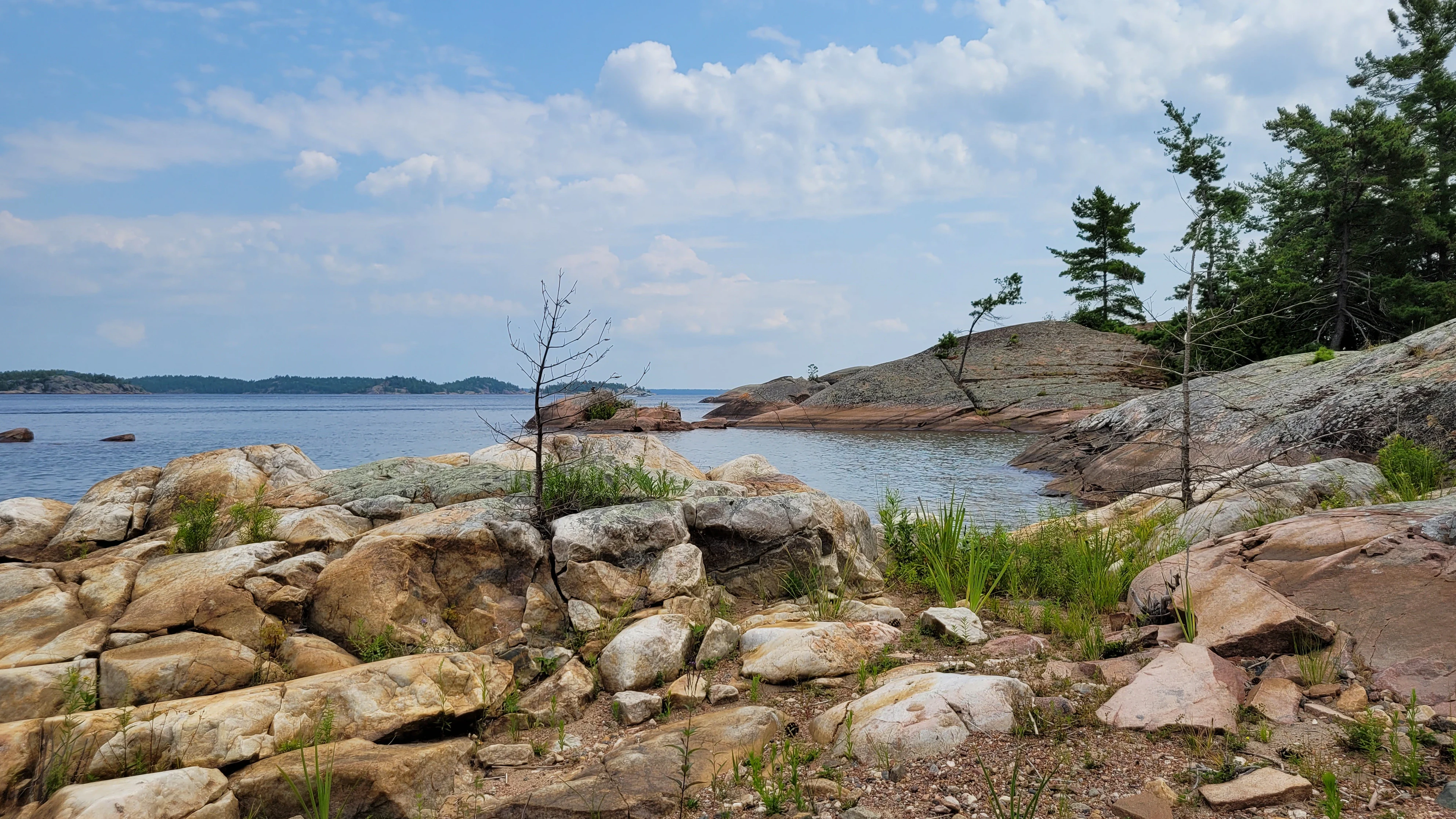 A Sandy beach on North Benjamin Island in Lake Huron with a white quartzite rock in the foreground, and pine trees, smooth granite, water and islands in the background