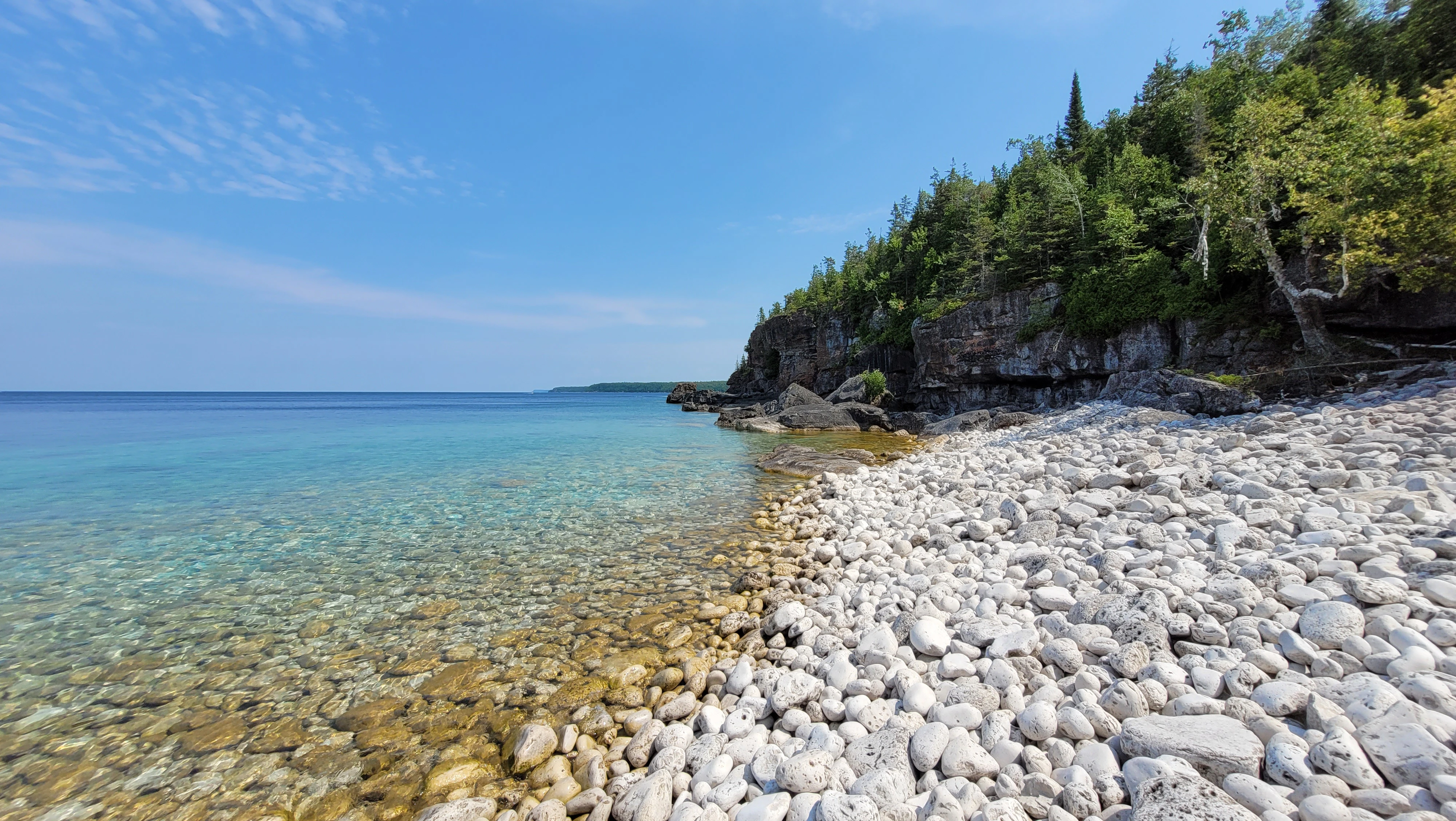 A white cobbelstone beach, clear turqoise water, cliffs and trees in the Bruce Peninsula in Ontario, Canada